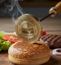 Burger press stamp on a hamburger bun with grill marks, surrounded by burger ingredients on a wooden board.