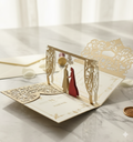 An intricate gold laser-cut wedding invitation card opened to reveal a 3D pop-up of a bride and groom silhouette standing under an archway. The card features "Walima" text and event details printed in gold on a cream background.
