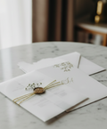A modern arch-shaped wedding invitation encased in a translucent vellum wrap. It is secured with a triple-wrapped gold string, a metallic gold wax seal, and a small sprig of dried white baby’s breath. A matching gold foil-stamped envelope sits in the background
