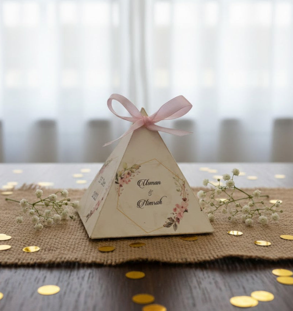 Triangular bidbox with floral design and pink ribbon on a table with confetti and flowers.