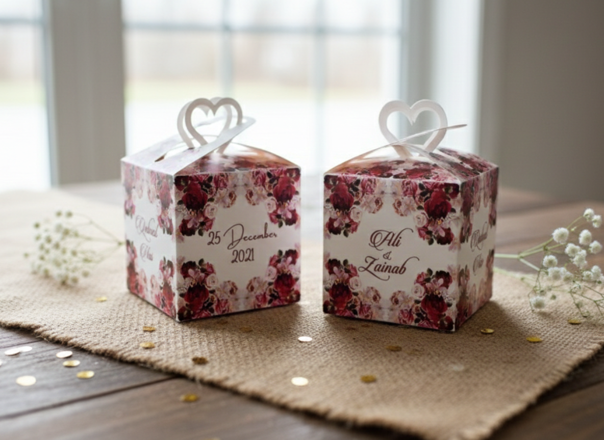 Two square wedding favor boxes with floral patterns, hearts as handles, and personalized text including dates and names.