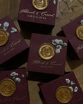 A collection of burgundy favor boxes with gold wax stamps, displaying names and dates, arranged on a surface.