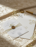 White invitation card with gold accents on a textured surface with light filtering through.
